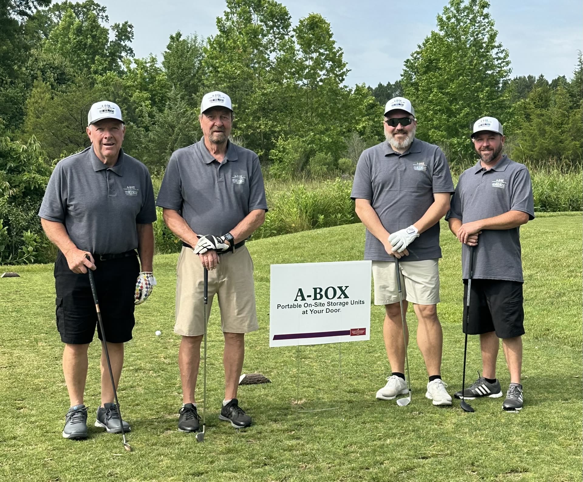 Four members of the A Box Portable Storage crew in branded shirts and caps beside an A Box sign at a local charity golf tournament