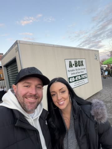 Two smiling A Box customers standing in front of an A Box portable storage container at a community event in Cambridge, Maryland