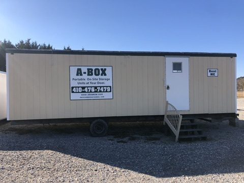 A tan A Box storage unit with an entry door and steps on the gravel rental lot in Cambridge, Maryland