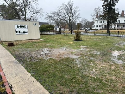 A Box storage container beside a fenced yard with a historic white farmhouse across the street in Cambridge, Maryland